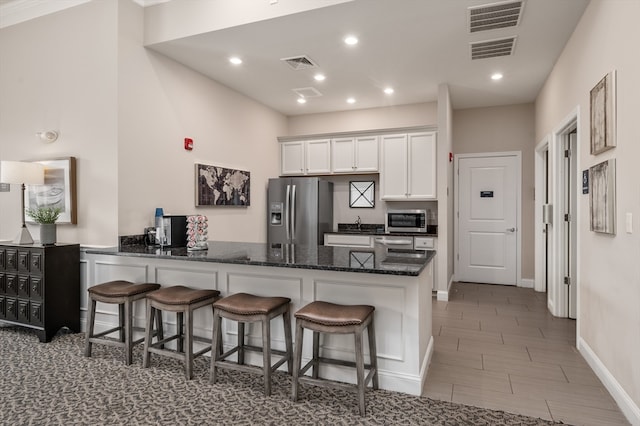 kitchen with kitchen peninsula, white cabinetry, stainless steel appliances, a kitchen breakfast bar, and dark stone countertops