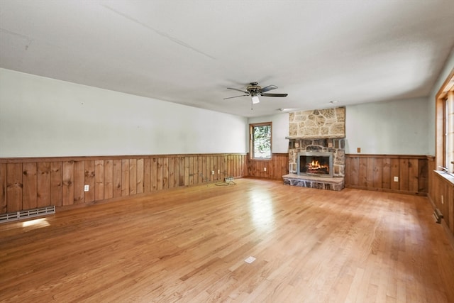 unfurnished living room featuring ceiling fan, wood walls, a stone fireplace, light hardwood / wood-style flooring, and a baseboard radiator