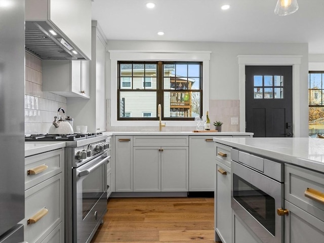 kitchen with backsplash, stainless steel appliances, wall chimney range hood, sink, and light hardwood / wood-style flooring