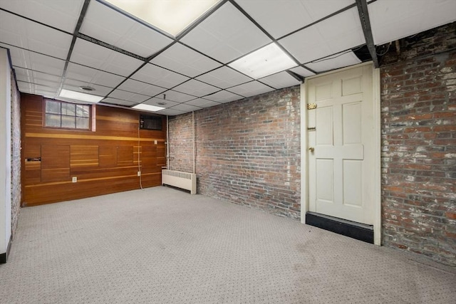 basement with a paneled ceiling, radiator heating unit, wooden walls, carpet flooring, and brick wall