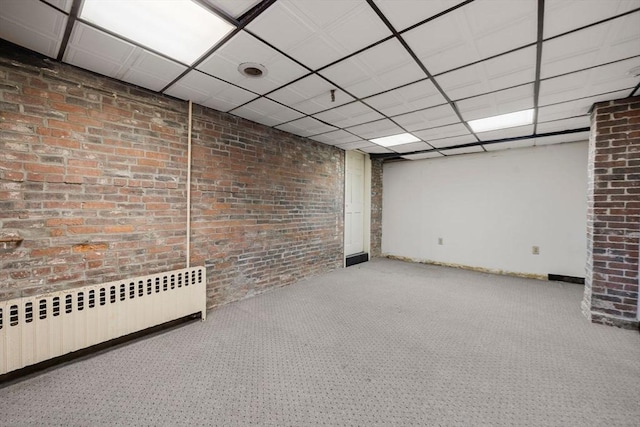 basement featuring brick wall, radiator heating unit, a drop ceiling, and carpet