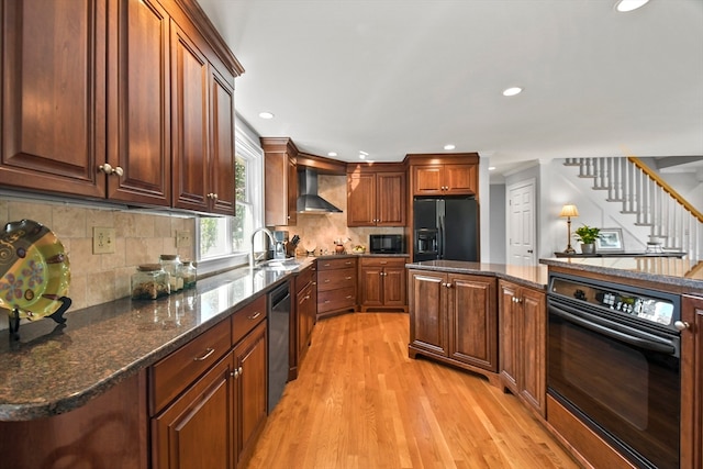 kitchen featuring wall chimney range hood, light wood-type flooring, a kitchen island, sink, and black appliances