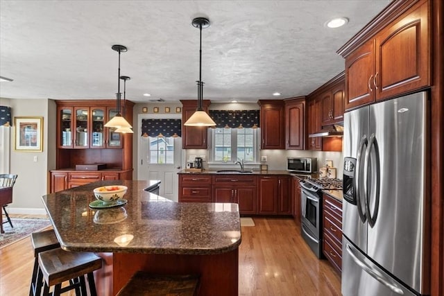 kitchen with appliances with stainless steel finishes, light wood-type flooring, a sink, glass insert cabinets, and under cabinet range hood