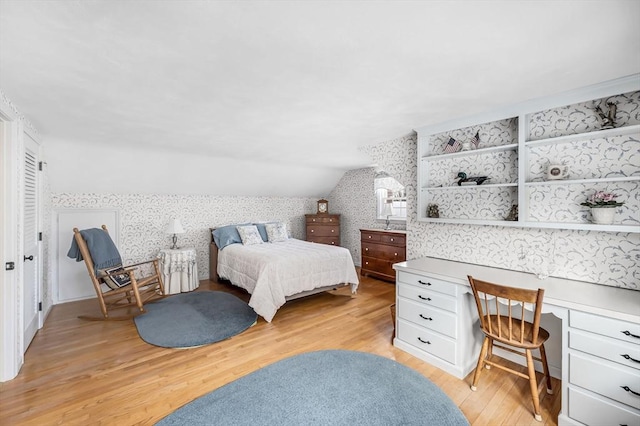 bedroom with light wood finished floors, built in desk, vaulted ceiling, and wallpapered walls