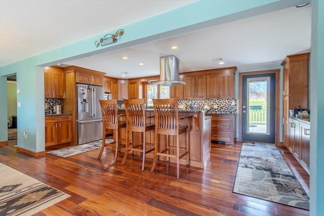 kitchen featuring island range hood, tasteful backsplash, brown cabinets, high quality fridge, and dark wood-type flooring