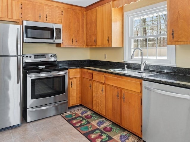 kitchen with appliances with stainless steel finishes, brown cabinetry, a sink, and dark stone counters