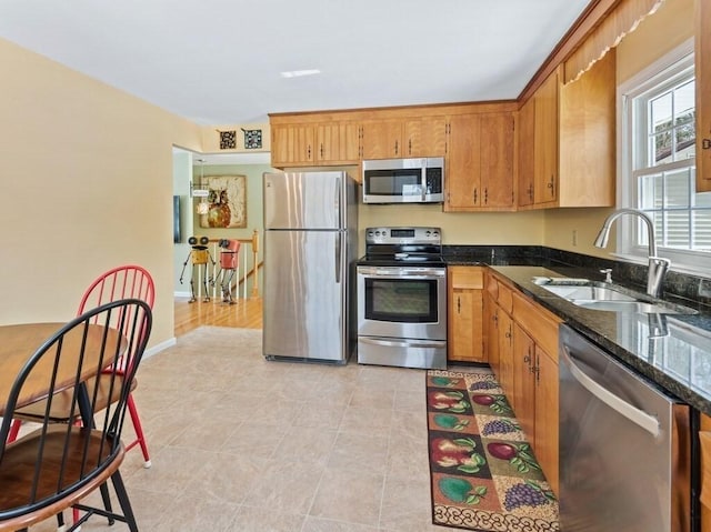 kitchen featuring baseboards, brown cabinetry, dark stone counters, appliances with stainless steel finishes, and a sink