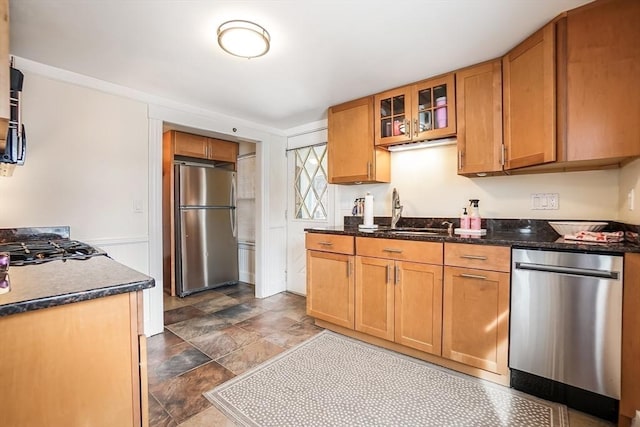 kitchen with a sink, appliances with stainless steel finishes, brown cabinetry, dark stone countertops, and glass insert cabinets