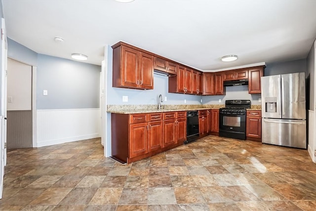 kitchen with wainscoting, stone finish flooring, under cabinet range hood, black appliances, and a sink