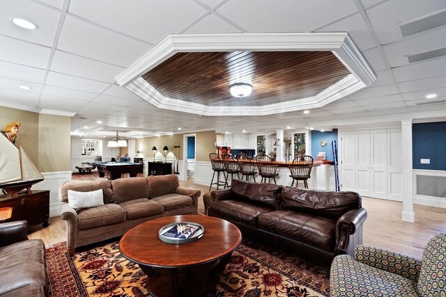 living room featuring indoor bar, ornamental molding, a tray ceiling, light wood-type flooring, and ornate columns