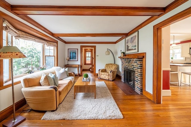 living room featuring a brick fireplace, beam ceiling, baseboards, light wood-style floors, and ornamental molding