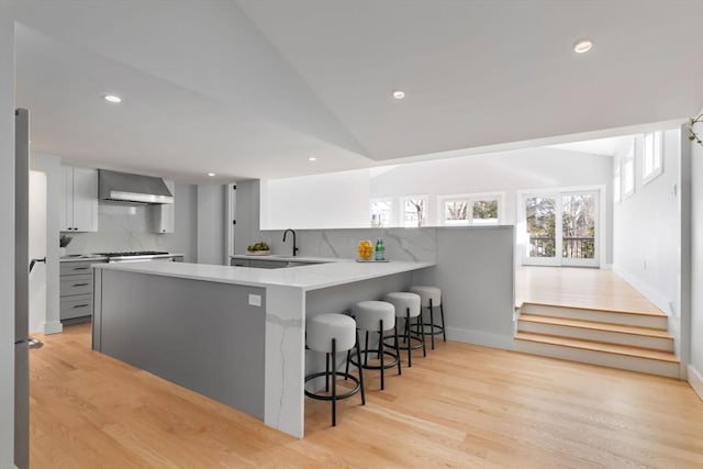 kitchen featuring a breakfast bar area, gray cabinetry, kitchen peninsula, decorative backsplash, and wall chimney range hood