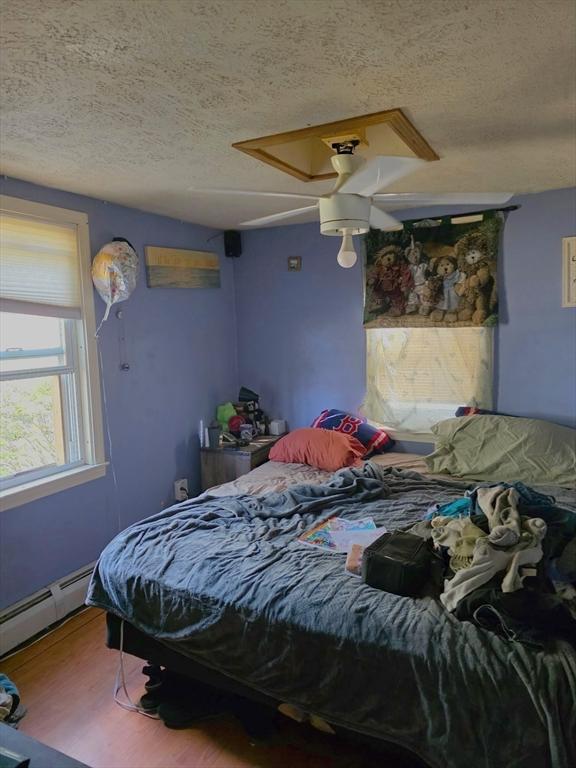 bedroom with a textured ceiling, baseboard heating, wood finished floors, and a ceiling fan