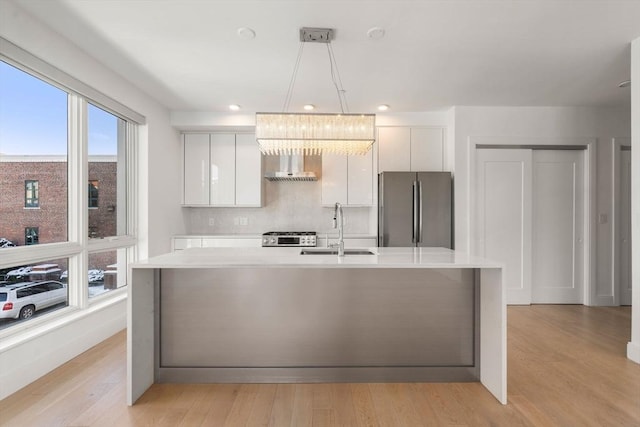 kitchen featuring wall chimney range hood, light countertops, white cabinets, appliances with stainless steel finishes, and a sink