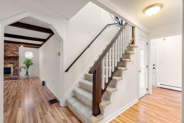 staircase with beamed ceiling, wood-type flooring, baseboard heating, and a brick fireplace