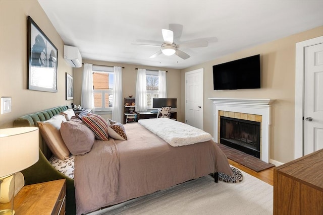 bedroom featuring a wall unit AC, wood finished floors, baseboards, ceiling fan, and a tile fireplace