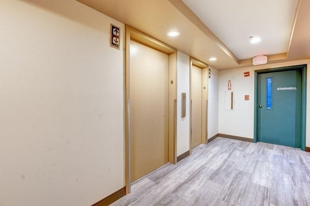corridor featuring light wood finished floors, a tray ceiling, elevator, recessed lighting, and baseboards