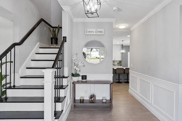 stairway with ornamental molding, a decorative wall, wood finished floors, a wainscoted wall, and a chandelier