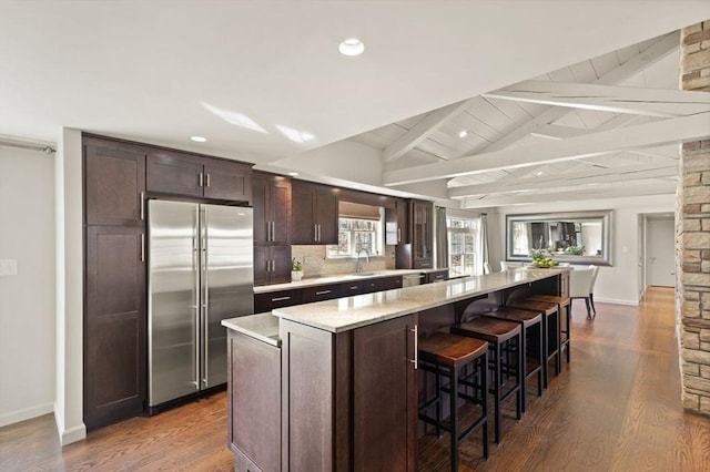 kitchen featuring dark brown cabinets, vaulted ceiling with beams, a breakfast bar, high quality fridge, and dark wood-style flooring