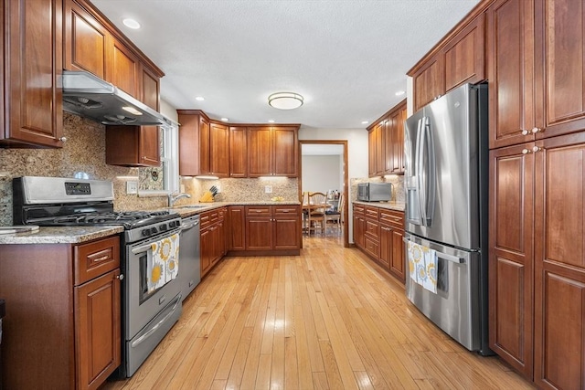kitchen with stainless steel appliances, under cabinet range hood, light wood-style flooring, decorative backsplash, and light stone counters