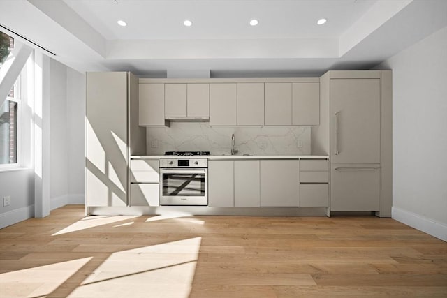 kitchen featuring tasteful backsplash, sink, a tray ceiling, light hardwood / wood-style flooring, and stainless steel oven