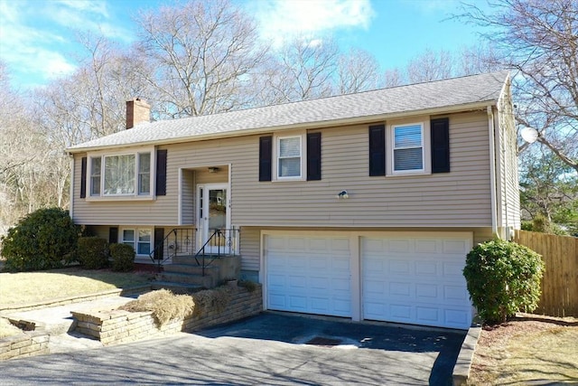 raised ranch with fence, driveway, an attached garage, a shingled roof, and a chimney