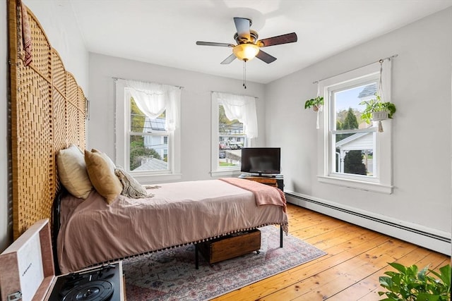 bedroom with a baseboard heating unit, multiple windows, wood-type flooring, and ceiling fan