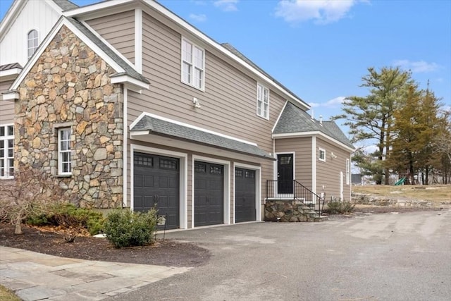 view of property exterior featuring a shingled roof, an attached garage, board and batten siding, stone siding, and driveway