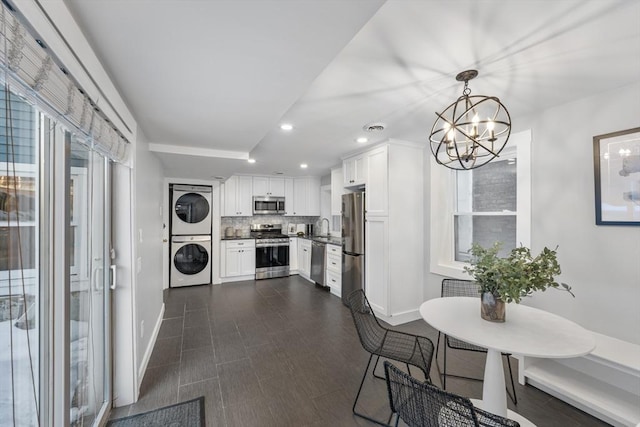 kitchen with appliances with stainless steel finishes, stacked washer / drying machine, hanging light fixtures, white cabinetry, and a sink