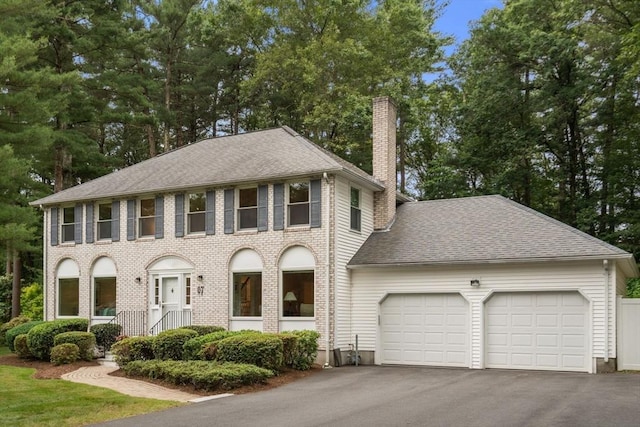 67 Flanagan Dr, Framingham, MA 01701 - colonial-style house featuring a chimney, an attached garage, driveway, roof with shingles, and brick siding