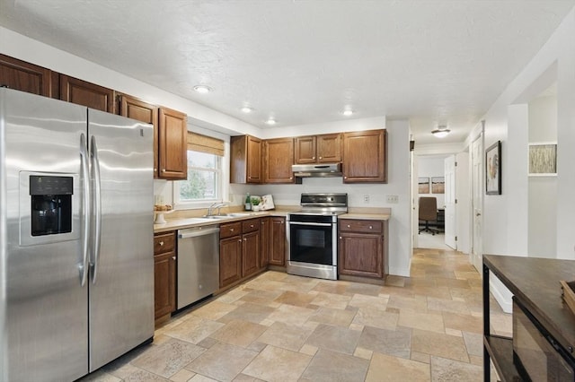 kitchen with light countertops, appliances with stainless steel finishes, stone finish flooring, a sink, and under cabinet range hood