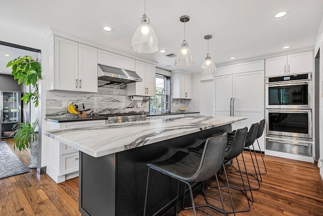 kitchen with tasteful backsplash, wall chimney range hood, double oven, dark wood-style floors, and white cabinetry