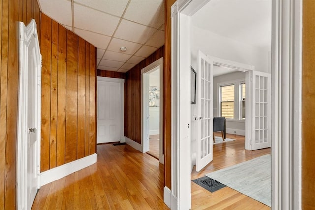 corridor featuring french doors, a paneled ceiling, baseboards, wooden walls, and light wood-style flooring