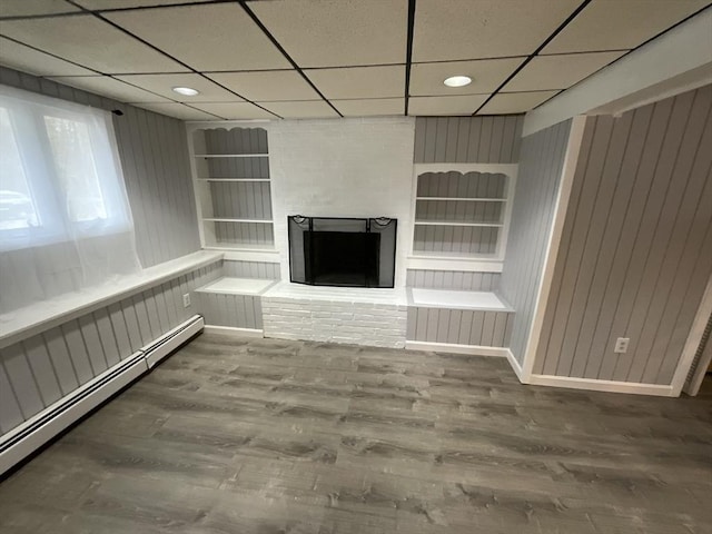 unfurnished living room with baseboard heating, a brick fireplace, a drop ceiling, and wood-type flooring