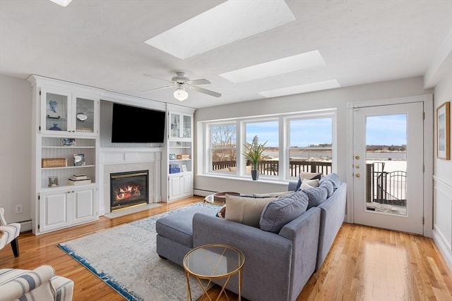 living area with a skylight, a fireplace with flush hearth, a ceiling fan, and light wood-style floors