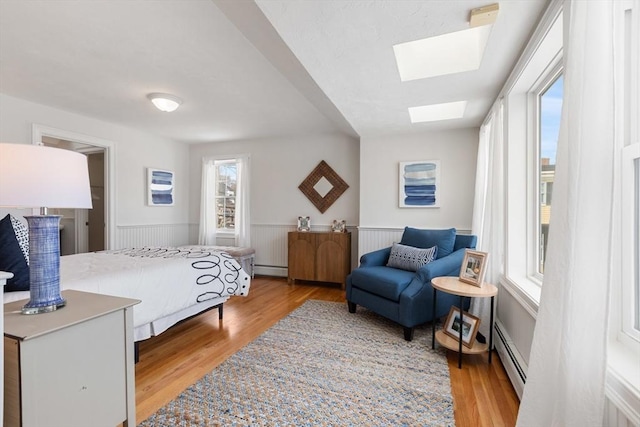 bedroom featuring a skylight, baseboard heating, light wood-style floors, and a wainscoted wall