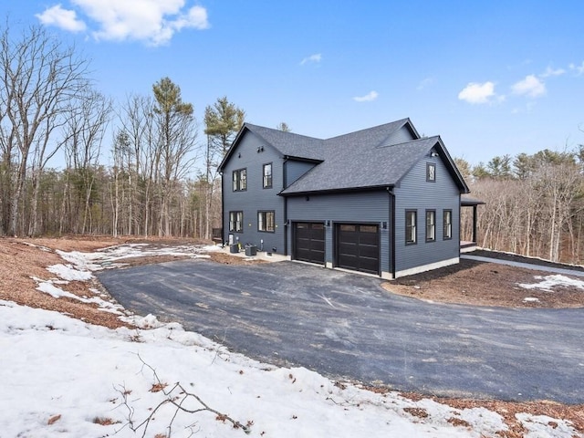 snow covered property with an attached garage, driveway, and a shingled roof