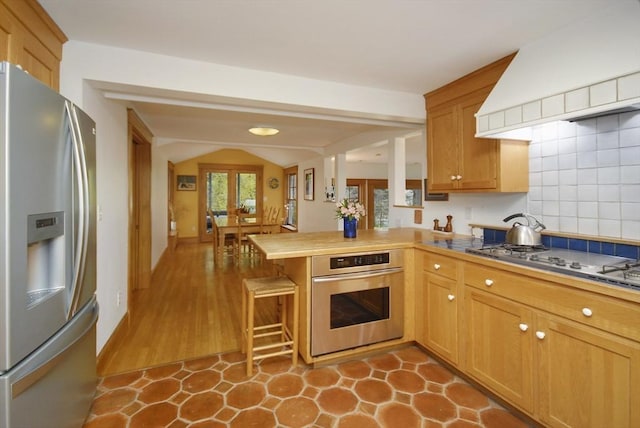 kitchen featuring a peninsula, a kitchen bar, custom range hood, appliances with stainless steel finishes, and vaulted ceiling