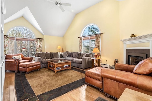 living area with a baseboard heating unit, a glass covered fireplace, ceiling fan, high vaulted ceiling, and hardwood / wood-style flooring