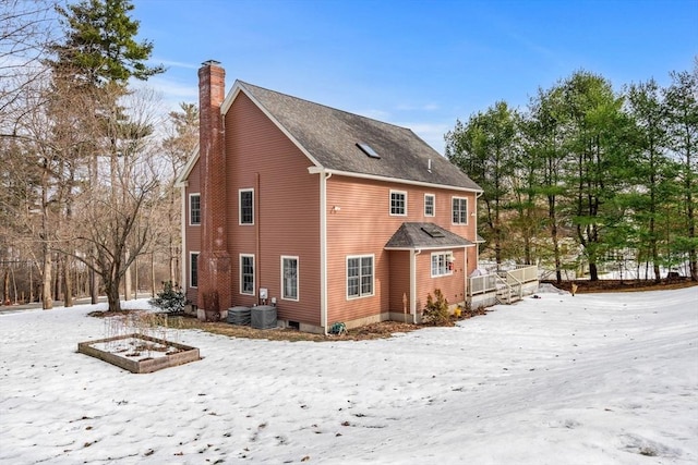 snow covered house with a shingled roof, cooling unit, and a chimney