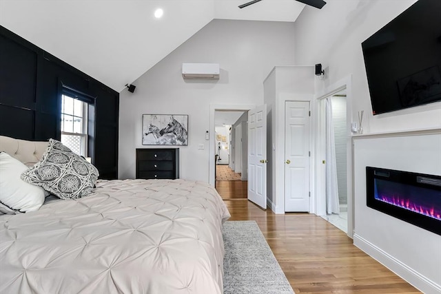 bedroom featuring baseboards, an AC wall unit, wood finished floors, a glass covered fireplace, and high vaulted ceiling