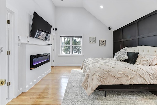 bedroom with a glass covered fireplace, baseboards, high vaulted ceiling, and light wood-style flooring