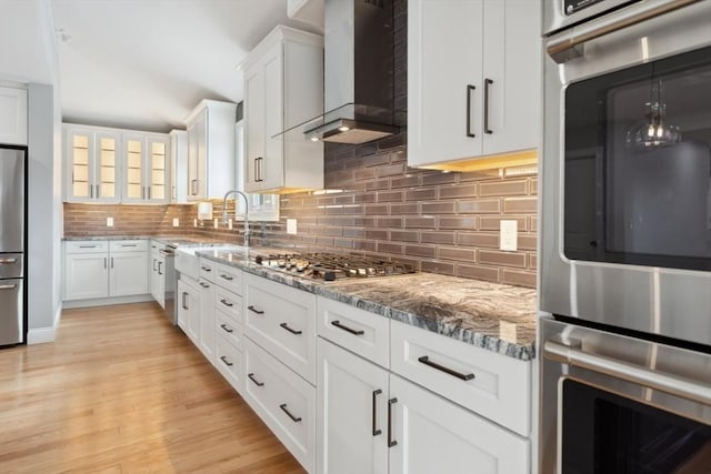 kitchen with stainless steel appliances, white cabinetry, wall chimney range hood, backsplash, and light stone countertops