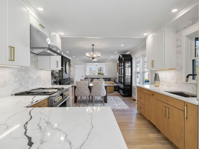 kitchen with wall chimney range hood, light stone countertops, sink, and white cabinets