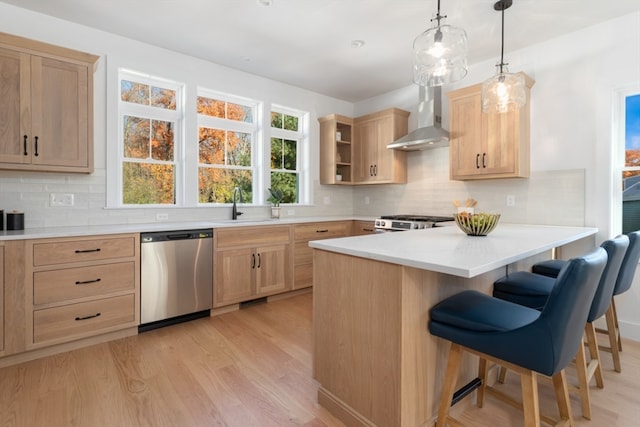 kitchen with wall chimney range hood, light brown cabinets, a breakfast bar area, appliances with stainless steel finishes, and pendant lighting