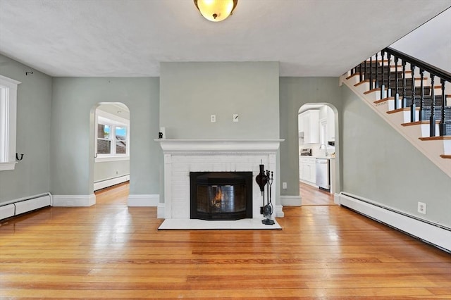 unfurnished living room featuring arched walkways, light wood finished floors, a baseboard heating unit, and stairs