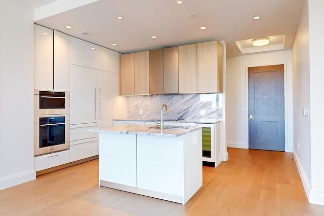 kitchen with sink, light stone counters, light wood-type flooring, stainless steel double oven, and a center island with sink