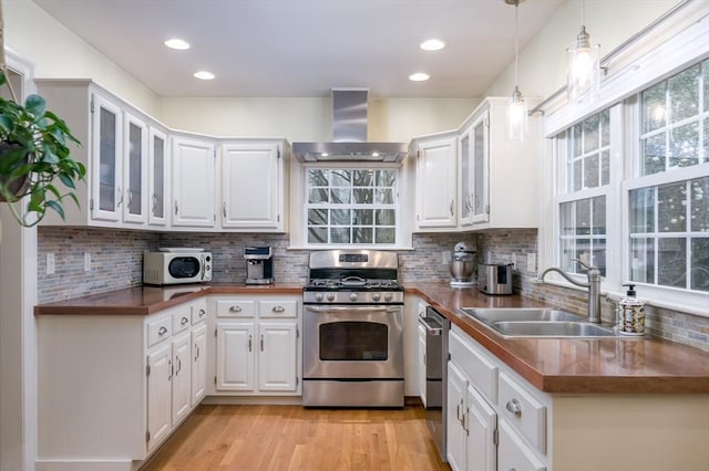 kitchen with stainless steel appliances, wall chimney range hood, a sink, white cabinetry, and glass insert cabinets