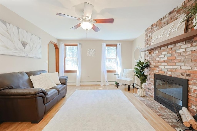 living room with a brick fireplace, light wood-style flooring, a baseboard radiator, and ceiling fan