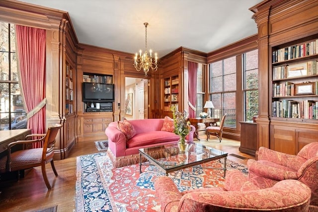 living area featuring an inviting chandelier, wooden walls, built in shelves, ornamental molding, and wood-type flooring